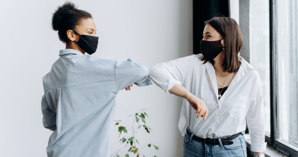 Two women in masks protect themselves from the COVID-19 pandemic by bumping elbows.