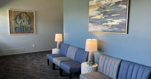 An interior photo of an Ellie Mental Health Therapy Clinic lobby with blue chairs, small reading lamps, and an Ellie Mental Health logo on one wall.