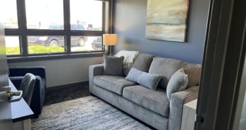 An interior photo of an Ellie Mental Health Therapy Clinic office with large windows, a gray couch, blue chair, small desk with office chair, and an abstract painting hanging on one wall.