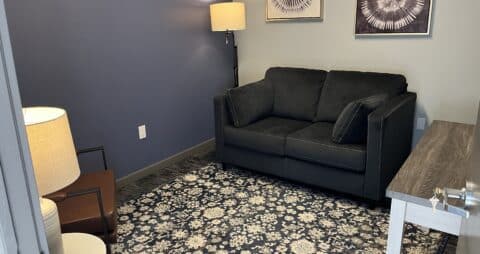 An interior photo of an Ellie Mental Health Therapy Clinic office that has floral patterned carpet, a dark love seat facing a brown leather chair, two lamps and two art pieces hanging on one wall.