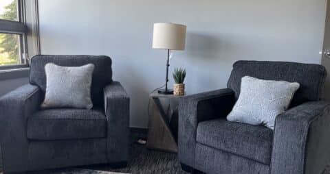 An interior photo of an Ellie Mental Health Therapy Clinic office with two dark gray chairs, a small table and table lamp.