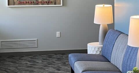 An interior photo of an Ellie Mental Health Therapy Clinic lobby with blue and gray chairs, a small white table with table lamps, and the Ellie Mental Health logo hanging on one wall.