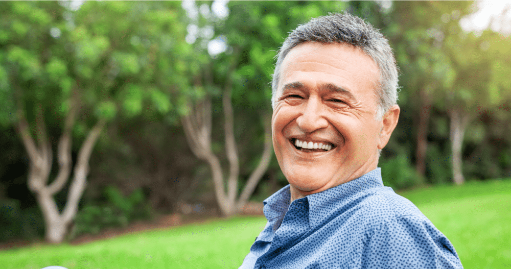 An older man sitting outside on the grass in a blue button up shirt smiling