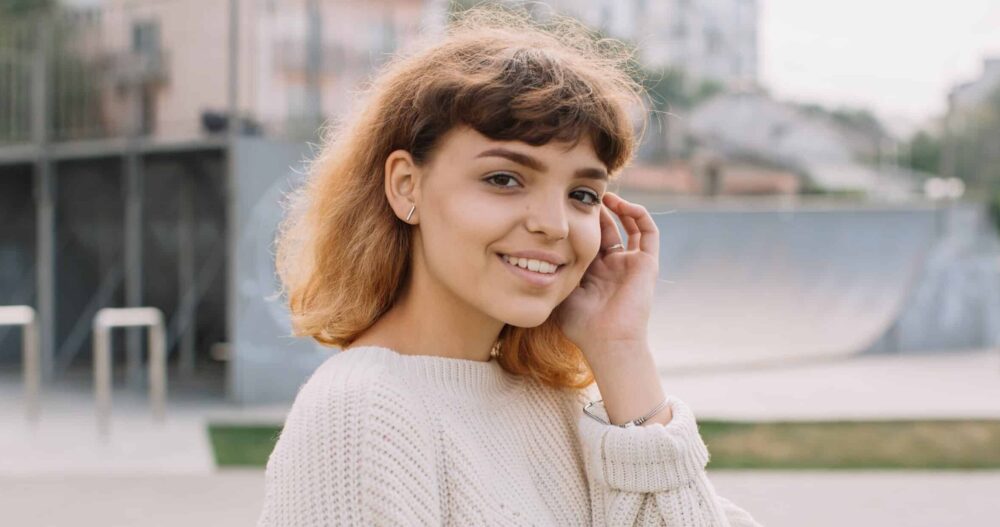 Woman standing outside smiling and brushing her hair behind her ear.