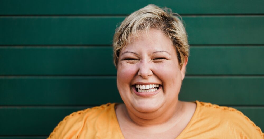 Blonde woman with short hair and an orange shirt standing in front of a green wall smiling