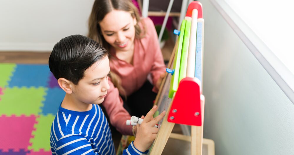 Child wearing a blue striped shirt drawing with his teacher next to him looking at his art and smiling