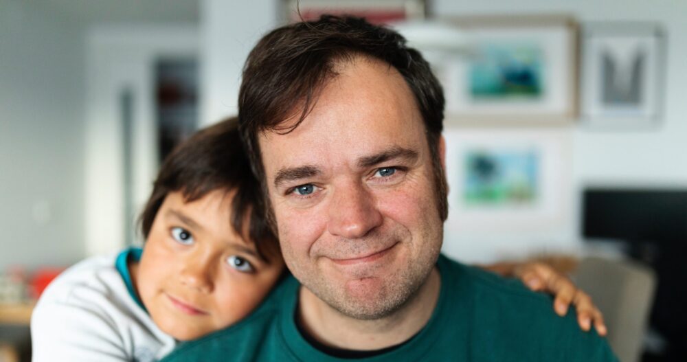 Man in a green shirt smiling with his toddler son hugging his right shoulder