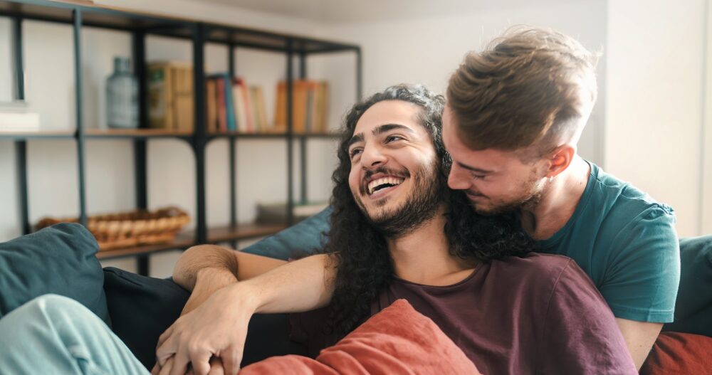 Heartwarming scene of two young gay men laughing and embracing on a sofa, showcasing love and affection.