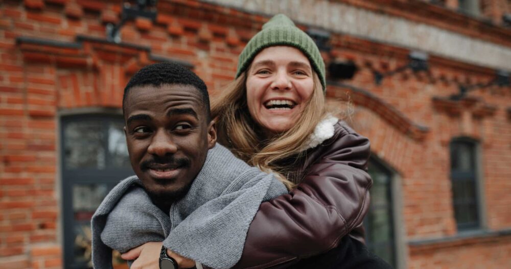 Happy woman riding on her male friend back on the street. Both dressed warmly for late autumn. Both having a great time, smiling