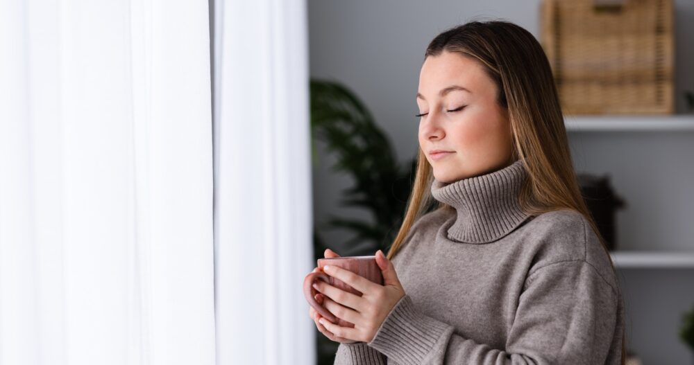 Winter clothing young woman breathing deep beside window while holding cup of hot drink as tea or coffee.