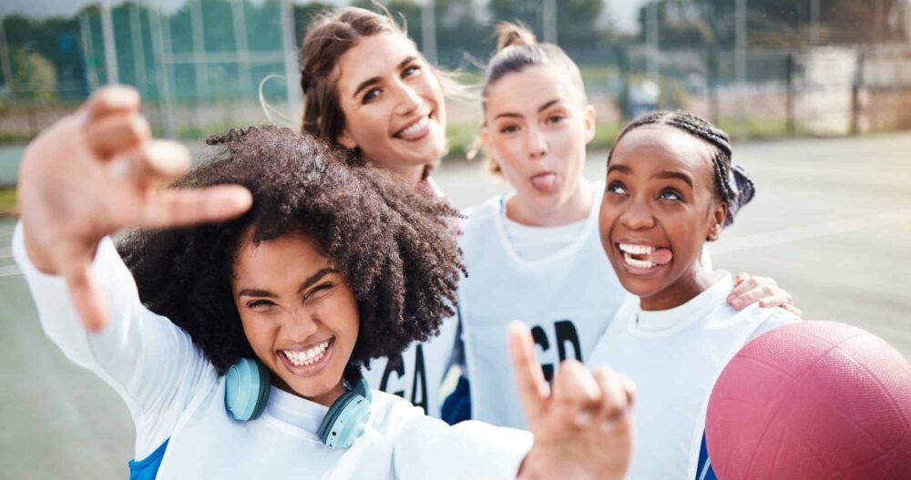 A group of girls holding a dodgeball pn thw blacktiop at recess. They are looking at the camera and smiling