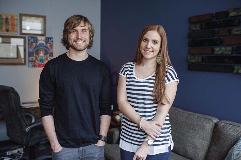 Ellie Mental Health founders Erin Pash, dressed in a white and blue striped shirt, and Kyle Keller, dressed in a dark blue shirt, standing next to each other inside one of the Ellie Mental Health offices.
