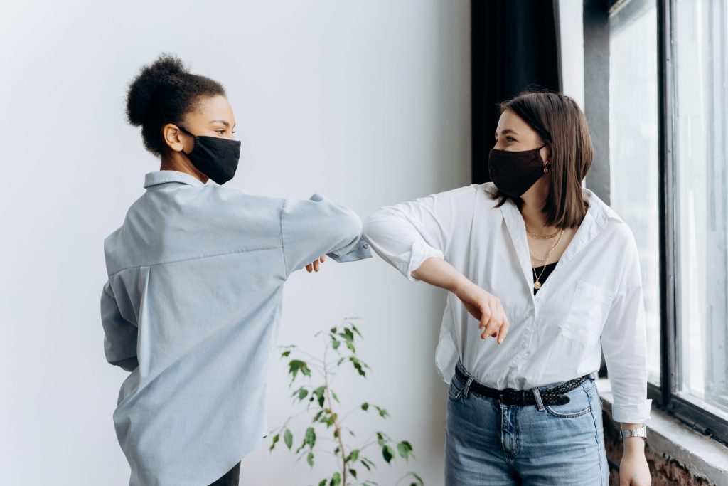 Two women in masks protect themselves from the COVID-19 pandemic by bumping elbows.