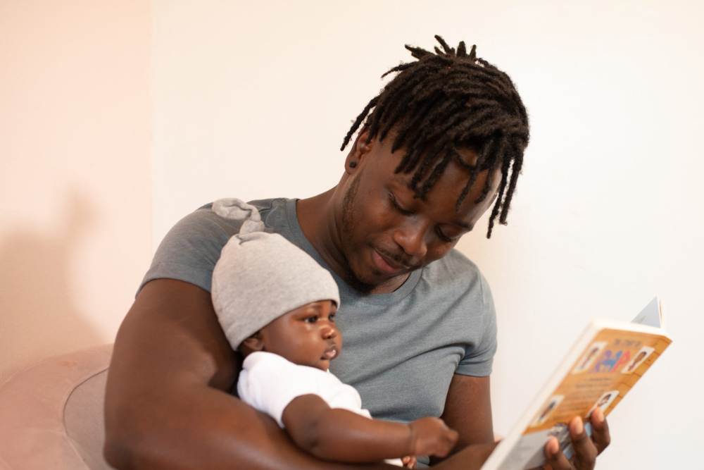 Man holding his young child and smiling at them while holding a book that he is reading to them