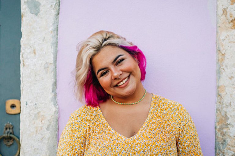 Woman with pink hair standing in front of a colorful wall and smiling