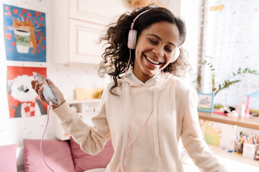 Happy teen in headphones listening to music and dancing in her cozy room at home