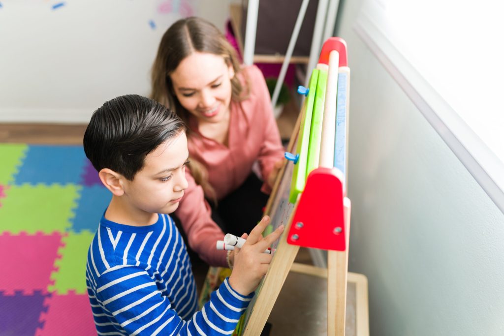 Child wearing a blue striped shirt drawing with his teacher next to him looking at his art and smiling