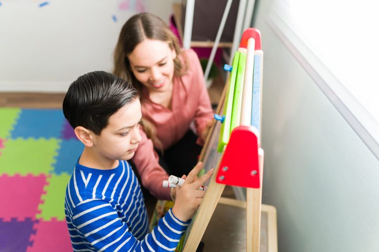 Child wearing a blue striped shirt drawing with his teacher next to him looking at his art and smiling