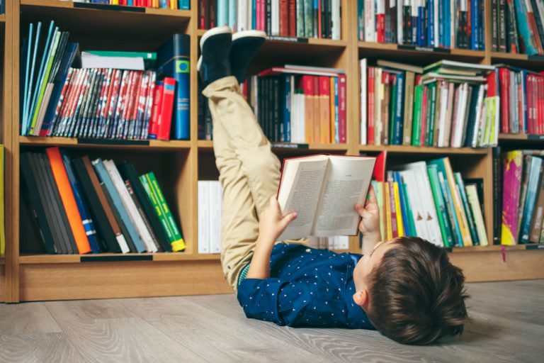 Boy laying on the floor with the feet up, reading a book against multi colored bookshelf in library.