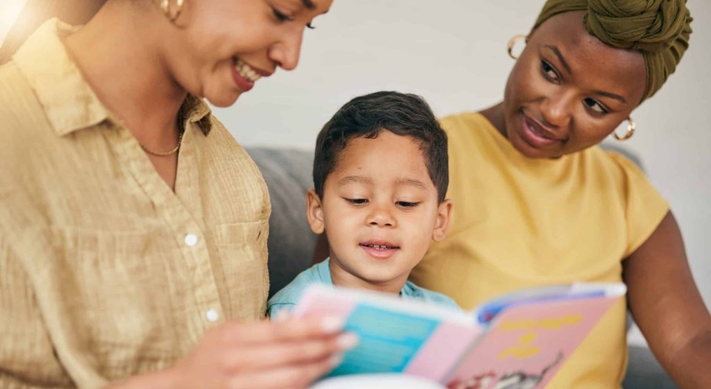 Two moms sitting on the couch with their young child reading a children's book
