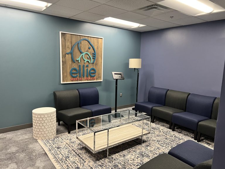 An interior photo of an Ellie Mental Health Therapy Clinic lobby with dark blue and gray chairs, a check-in kiosk, a small white table and a larger glass table, a floor lamp, and the Ellie Mental Health logo hanging on one wall.