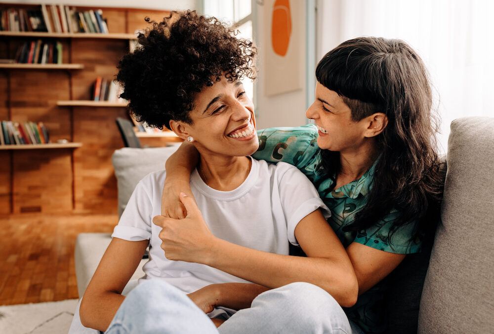 Happy young lesbian couple sharing a laugh while sitting together in their living room.