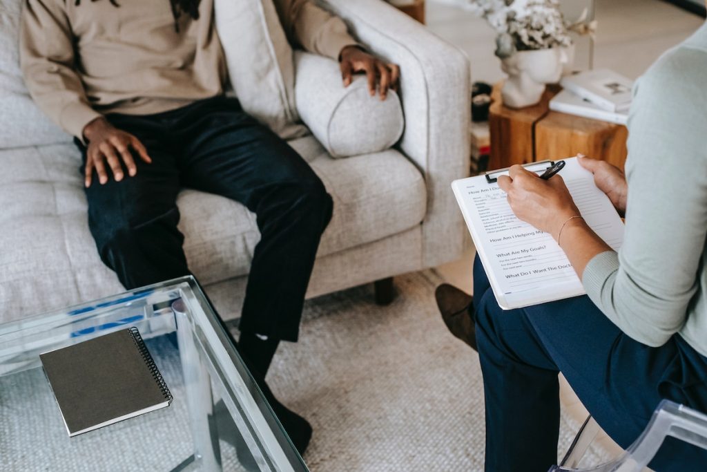 Black man sitting on a cream colored couch across from a woman therapist holding a clipboard taking notes