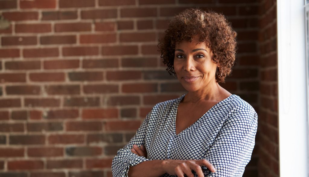 A black woman wearing a white and blue striped shirt standing in front of a brick wall with her arms crossed