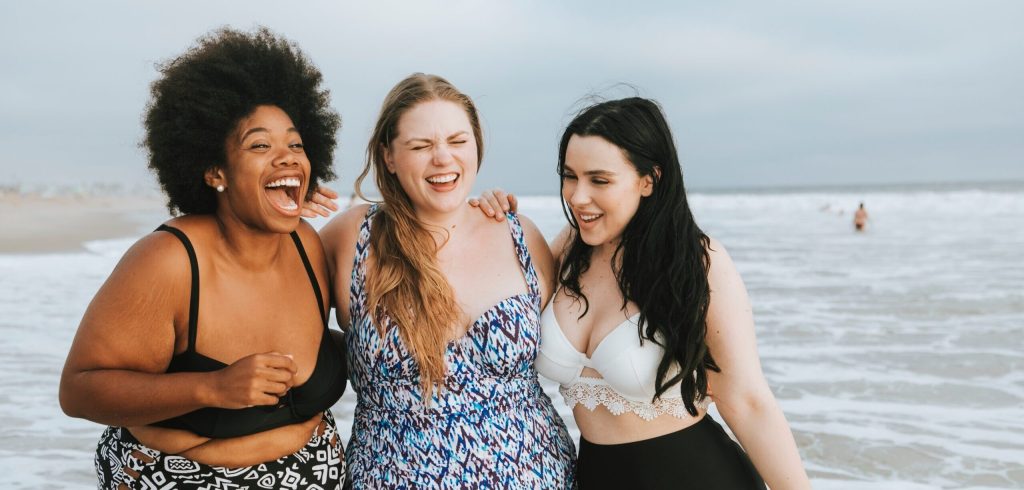Three women on the beach with the ocean behind them. They are hugging and smiling inf ront of the water