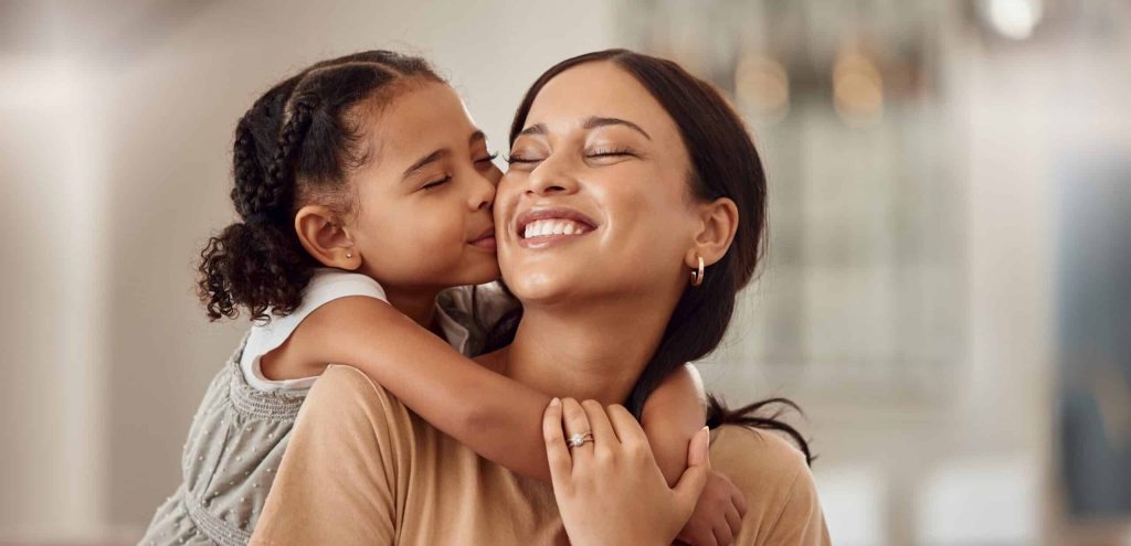 Child hugging her mom from behind and the mom turning her head and smiling at her