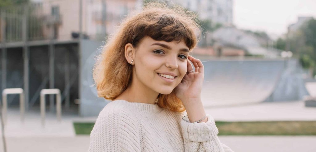 Woman standing outside smiling and brushing her hair behind her ear.