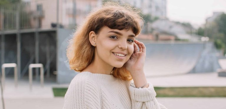 Woman standing outside smiling and brushing her hair behind her ear.