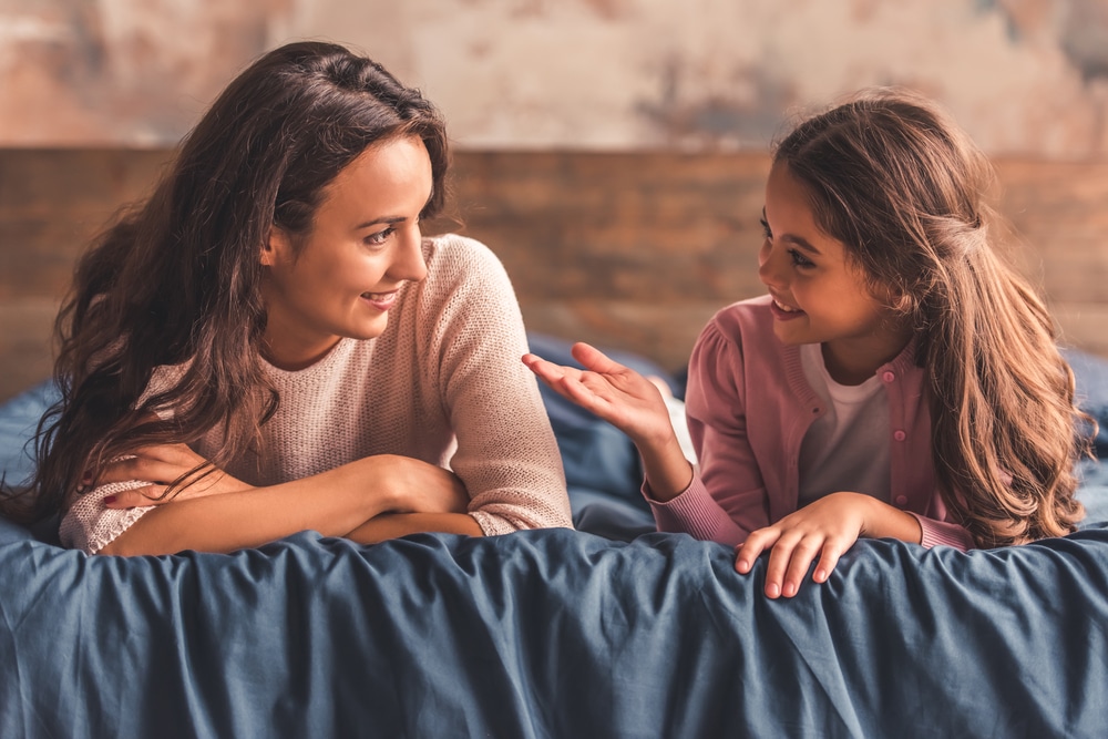 A mom and daughter talk about mental health while lying on a bed.