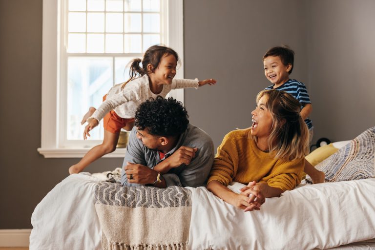 Happy family laughing and having fun together on the bed. Two parents playing with their son and daughter at home. Mom and dad spending quality time with their children on the weekend.